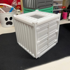 May include: A white miniature shipping container with a hinged door and a small opening on the top. The container is made of plastic and has a realistic design.