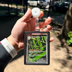 K&ouml;nnte beinhalten: Ein schwarzer Kartenhalter mit einer einziehbaren Rolle, der eine Pok&eacute;mon-Sammelkarte h&auml;lt. Die Karte hat ein gr&uuml;nes und schwarzes Design mit dem Text "Toxapex Spidops". Der Halter wird in einer Hand gehalten.