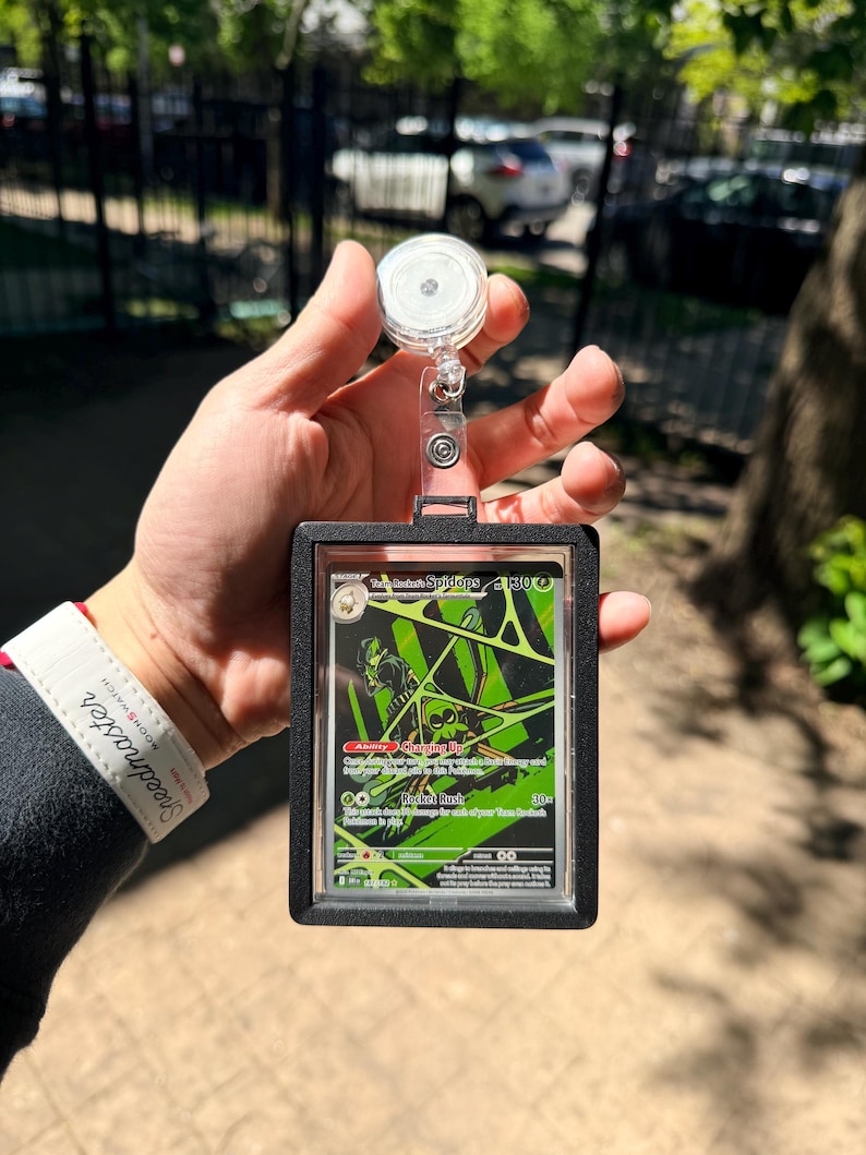 K&ouml;nnte beinhalten: Ein schwarzer Kartenhalter mit einer einziehbaren Rolle, der eine Pok&eacute;mon-Sammelkarte h&auml;lt. Die Karte hat ein gr&uuml;nes und schwarzes Design mit dem Text "Toxapex Spidops". Der Halter wird in einer Hand gehalten.