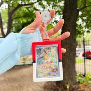May include: A red, white, and black plastic card holder frame designed to resemble a Poké Ball, containing a trading card featuring a sleeping Eevee character. The frame is attached to a clear, circular retractable badge reel with a metal clip.