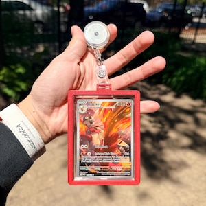 K&ouml;nnte beinhalten: Ein roter Ausweishalter mit einer einziehbaren Rolle, der eine Pok&eacute;mon-Sammelkarte h&auml;lt. Die Karte zeigt eine feurige Illustration von Lohgock mit Text wie "Heat Blast" und "Inferno Kick Fury".