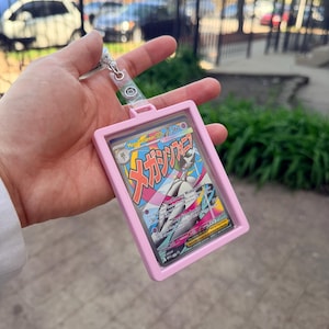 K&ouml;nnte beinhalten: Ein rosa Kartenhalter mit einem transparenten Kunststofffenster, der eine farbenfrohe Sammelkarte enth&auml;lt. Die Karte enth&auml;lt japanischen Text und eine Charakterillustration. Eine silberfarbene Kette und ein Verschluss sind am Halter befestigt.