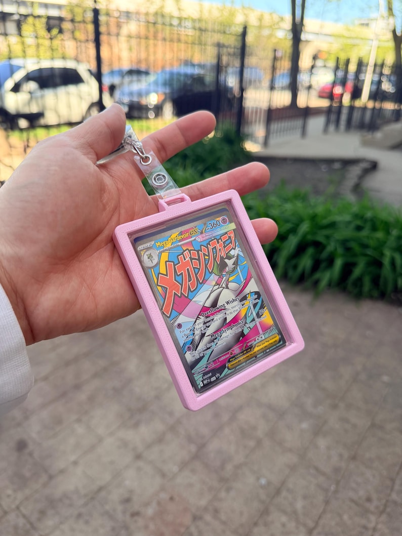 K&ouml;nnte beinhalten: Ein rosa Kartenhalter mit einem transparenten Kunststofffenster, der eine farbenfrohe Sammelkarte enth&auml;lt. Die Karte enth&auml;lt japanischen Text und eine Charakterillustration. Eine silberfarbene Kette und ein Verschluss sind am Halter befestigt.