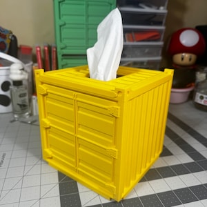May include: A yellow tissue box holder shaped like a shipping container. The tissue box is partially pulled out of the top opening.
