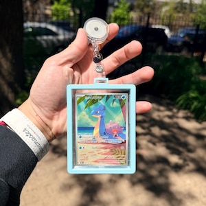 K&ouml;nnte beinhalten: Ein hellblauer Ausweishalter mit einer einziehbaren Rolle, der eine Pok&eacute;mon-Sammelkarte mit Lapras enth&auml;lt. Die Karte zeigt Lapras in einer tropischen Umgebung mit dem Text "Swim Together" und "Surf".