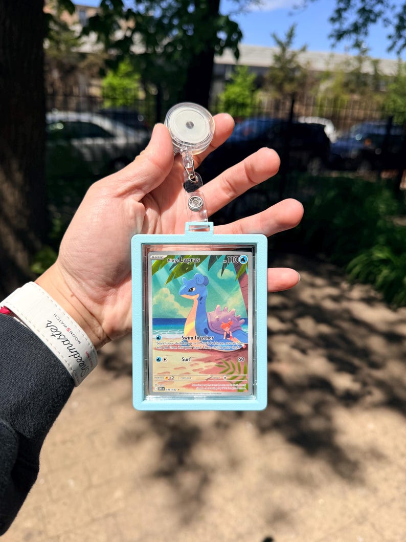 K&ouml;nnte beinhalten: Ein hellblauer Ausweishalter mit einer einziehbaren Rolle, der eine Pok&eacute;mon-Sammelkarte mit Lapras enth&auml;lt. Die Karte zeigt Lapras in einer tropischen Umgebung mit dem Text "Swim Together" und "Surf".