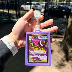 K&ouml;nnte beinhalten: Ein lila Ausweishalter mit einem transparenten Kunststofffenster, der eine Pok&eacute;mon-Karte mit Mega Gengar GX enth&auml;lt. Die Karte hat japanischen Text und farbenfrohe Grafiken. Der Halter ist an einer einziehbaren Rolle mit einem transparenten Kunststoffclip befestigt.