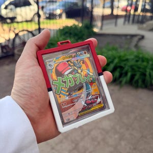 K&ouml;nnte beinhalten: Ein roter, wei&szlig;er und klarer Kunststoff-Kartenhalter mit einer Pok&eacute;mon-Sammelkarte. Die Karte zeigt eine farbenfrohe Illustration eines Mega-Lucario-Charakters mit japanischem Text. Der Halter ist rechteckig mit einer kleinen Schlaufe oben.
