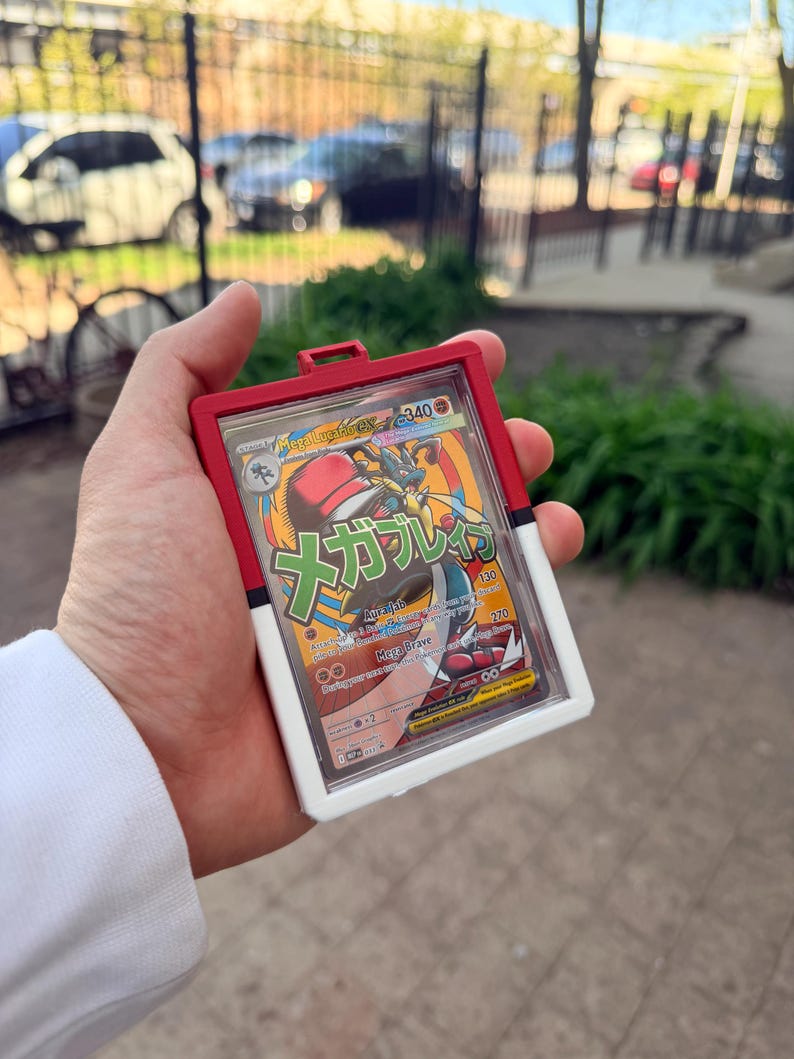K&ouml;nnte beinhalten: Ein roter, wei&szlig;er und klarer Kunststoff-Kartenhalter mit einer Pok&eacute;mon-Sammelkarte. Die Karte zeigt eine farbenfrohe Illustration eines Mega-Lucario-Charakters mit japanischem Text. Der Halter ist rechteckig mit einer kleinen Schlaufe oben.