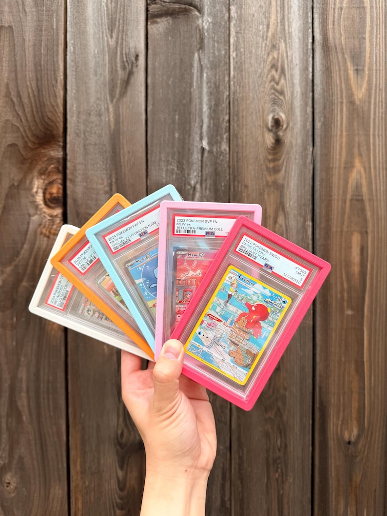May include: A collection of five graded trading cards in colorful plastic frames, including white, orange, light blue, pink, and magenta. Each card is encased in a protective slab with a PSA label at the top, displaying various Pok&eacute;mon illustrations.