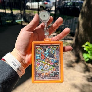 K&ouml;nnte beinhalten: Ein orangefarbener Ausweishalter mit einem transparenten Kunststofffenster, der eine farbenfrohe Sammelkarte mit der Figur Rotom enth&auml;lt. Die Karte hat eine lebendige Illustration mit dem Text "Rotom" und "Gadget Show". Eine einziehbare Ausweisrolle ist angebracht.