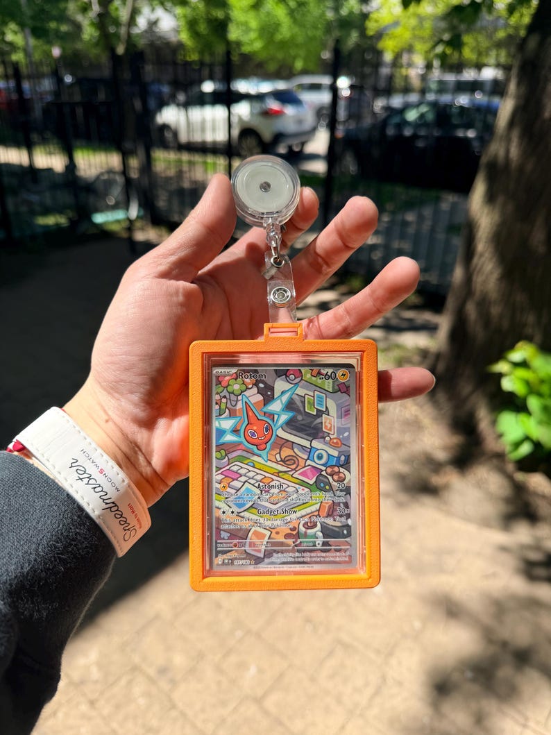 K&ouml;nnte beinhalten: Ein orangefarbener Ausweishalter mit einem transparenten Kunststofffenster, der eine farbenfrohe Sammelkarte mit der Figur Rotom enth&auml;lt. Die Karte hat eine lebendige Illustration mit dem Text "Rotom" und "Gadget Show". Eine einziehbare Ausweisrolle ist angebracht.