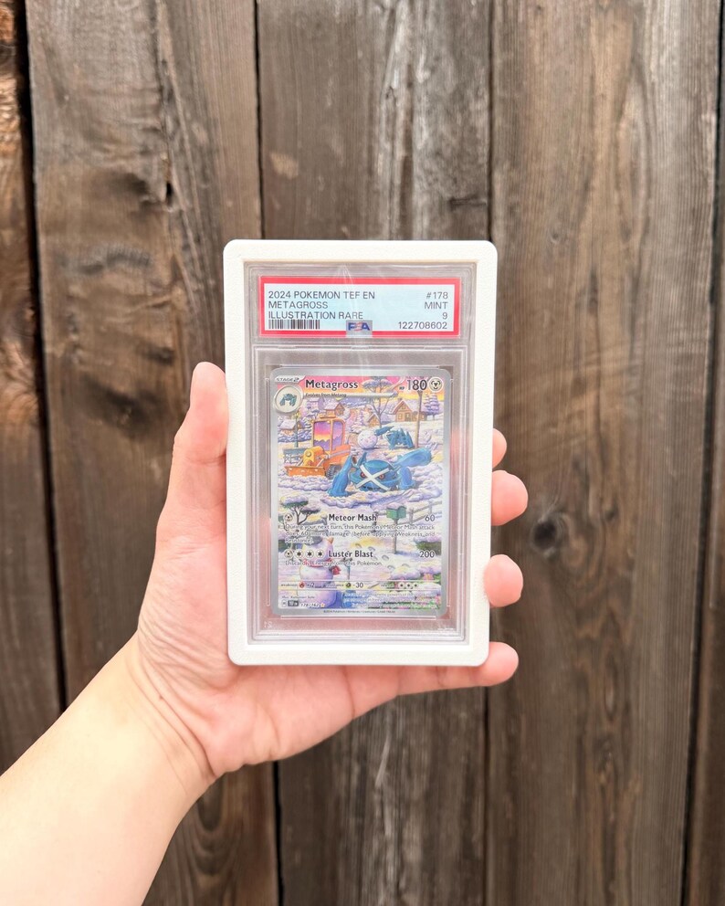 May include: A 2024 Pokemon Metagross Illustration Rare trading card, graded Mint 9 by PSA, encased in a white protective plastic slab. The card features a snowy landscape illustration with the character Metagross, Meteor Mash text, and 180 HP.