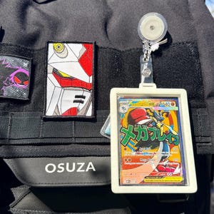 K&ouml;nnte beinhalten: Schwarzer taktischer Rucksack mit Aufn&auml;hern und einem Kartenhalter. Zu den Aufn&auml;hern geh&ouml;ren ein Robotergesicht und ein lila Geist. Der Kartenhalter zeigt eine Pok&eacute;mon-Karte mit japanischem Text und einer Lucario-Illustration. Auf dem Rucksack steht das Wort "OSUZA".