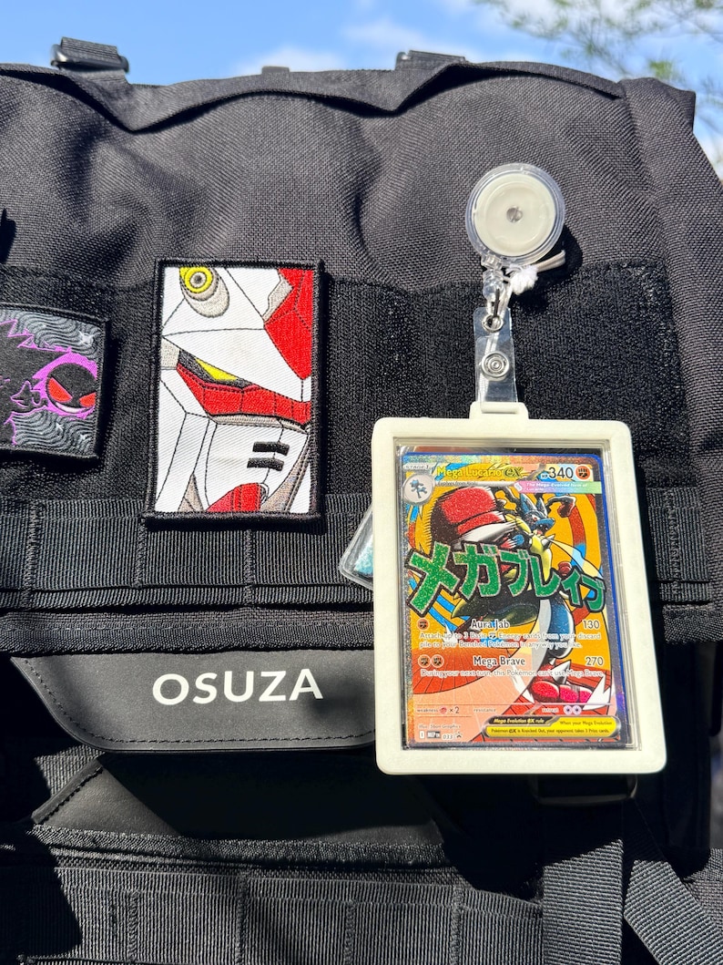 K&ouml;nnte beinhalten: Schwarzer taktischer Rucksack mit Aufn&auml;hern und einem Kartenhalter. Zu den Aufn&auml;hern geh&ouml;ren ein Robotergesicht und ein lila Geist. Der Kartenhalter zeigt eine Pok&eacute;mon-Karte mit japanischem Text und einer Lucario-Illustration. Auf dem Rucksack steht das Wort "OSUZA".