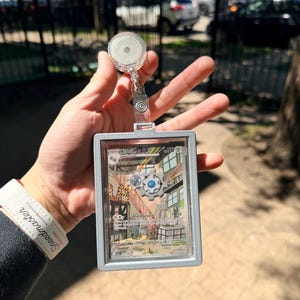 K&ouml;nnte beinhalten: Ein durchsichtiger, einziehbarer Ausweishalter aus Kunststoff mit einem silbernen Rahmen, der eine Sammelkarte h&auml;lt. Die Karte zeigt eine farbenfrohe Illustration einer mechanischen Szene mit dem Namen "Klang" und dem Text "Hard Gears". Der Ausweishalter wird vor einem verschwommenen Hintergrund im Freien gehalten.