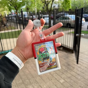 K&ouml;nnte beinhalten: Ein rot-wei&szlig;er Ausweishalter mit einziehbarem Aufroller, der eine Pok&eacute;mon-Sammelkarte h&auml;lt. Die Karte zeigt eine farbenfrohe Illustration mit japanischem Text. Der Ausweishalter wird vor einem verschwommenen Hintergrund im Freien gehalten.