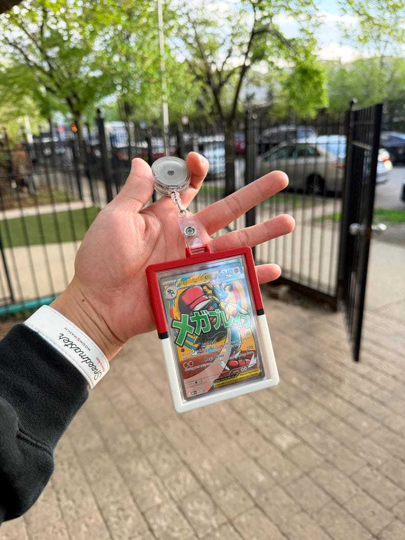 K&ouml;nnte beinhalten: Ein rot-wei&szlig;er Ausweishalter mit einziehbarem Aufroller, der eine Pok&eacute;mon-Sammelkarte h&auml;lt. Die Karte zeigt eine farbenfrohe Illustration mit japanischem Text. Der Ausweishalter wird vor einem verschwommenen Hintergrund im Freien gehalten.