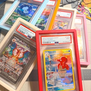 May include: A collection of five PSA-graded trading cards in colorful plastic cases, including a 2023 Charizard ex and a 2022 Octillery card. The cards are displayed in light blue, orange, cream, red, and pink frames on a grid-patterned surface.