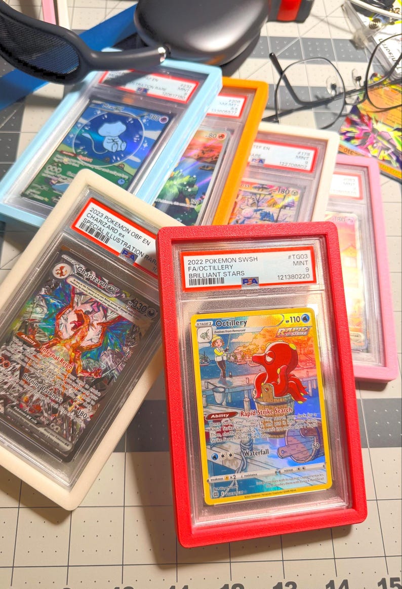May include: A collection of five PSA-graded trading cards in colorful plastic cases, including a 2023 Charizard ex and a 2022 Octillery card. The cards are displayed in light blue, orange, cream, red, and pink frames on a grid-patterned surface.