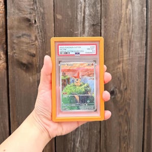 May include: A graded 2025 Pokemon SVP EN Victini trading card in a protective orange plastic slab. The card features an illustration of the character Victini on a grassy cliffside overlooking water, with the text Victini and 80 HP visible.