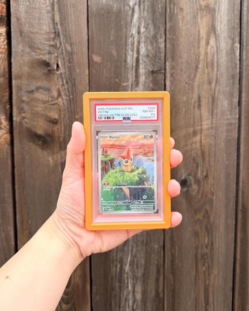 May include: A graded 2025 Pokemon SVP EN Victini trading card in a protective orange plastic slab. The card features an illustration of the character Victini on a grassy cliffside overlooking water, with the text Victini and 80 HP visible.