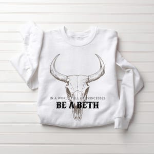 May include: White sweatshirt with a bull skull graphic. The text "IN A WORLD FULL OF PRINCESSES BE A BETH" is printed below the skull in black. The sweatshirt is laid flat on a white surface.