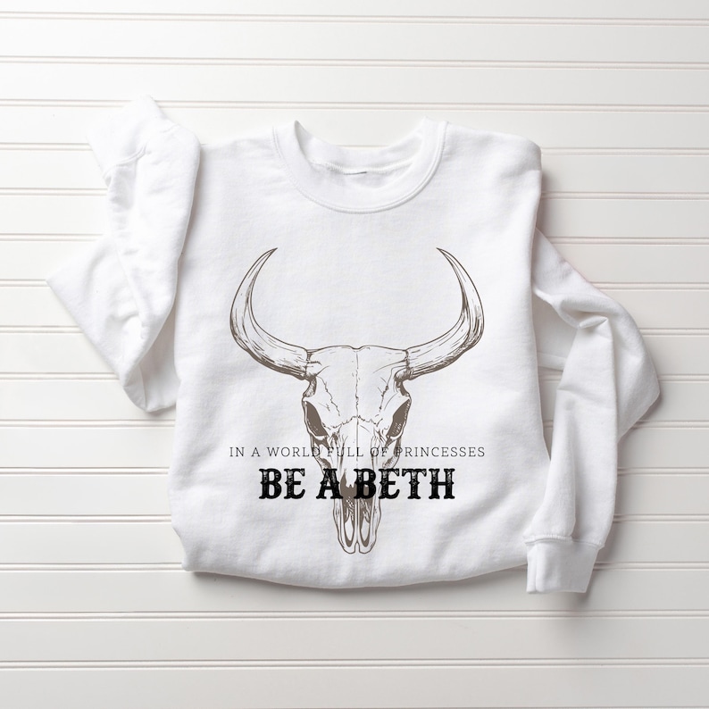 May include: White sweatshirt with a bull skull graphic. The text "IN A WORLD FULL OF PRINCESSES BE A BETH" is printed below the skull in black. The sweatshirt is laid flat on a white surface.