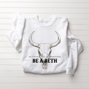 May include: White sweatshirt with a bull skull graphic. The text "IN A WORLD FULL OF PRINCESSES BE A BETH" is printed below the skull in black. The sweatshirt is laid flat on a white surface.
