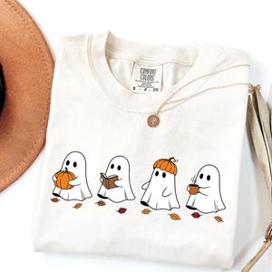 May include: An off-white t-shirt featuring a graphic of four white ghosts. One ghost holds an orange pumpkin, one reads a brown book, one wears a pumpkin as a hat, and one holds a brown mug. Small scattered autumn leaves are printed below the ghosts.