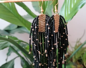May include: A pair of long fringe earrings featuring a rectangular top section woven with small metallic gold beads. Numerous strands of black seed beads hang from the base, accented with intermittent metallic gold beads throughout the length.