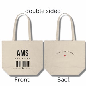 Peut inclure: Un sac fourre-tout en toile blanche avec du texte noir sur le devant qui dit "AMS AMSTERDAM" avec un code-barres et des coordonnées. Le dos du sac porte le texte "HOME IS AMSTERDAM" en arc de cercle avec un cœur rouge.