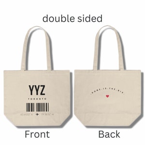 May include: A white canvas tote bag with a black and white airport style luggage tag on the front that reads "YYZ TORONTO" with a barcode and coordinates. The back of the bag has the text "HOME IS THE 6IX" with a red heart.
