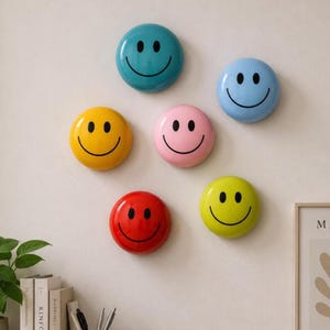 May include: A collection of six smiley face wall decorations in various colors: teal, light blue, yellow, pink, red, and lime green. Each circular decoration features a black smiling face. The decorations are mounted on a white wall.