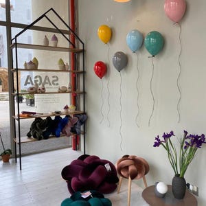 May include: A black metal house-shaped shelving unit displays ceramic cupcakes and knot pillows. The wall features six balloon-shaped wall lamps in red, yellow, gray, blue, teal, and pink. Two large velvet knot floor cushions and a small stool sit nearby.