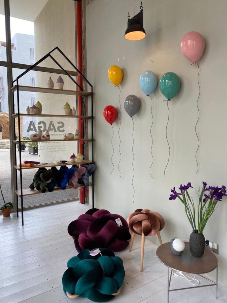 May include: A black metal house-shaped shelving unit displays ceramic cupcakes and knot pillows. The wall features six balloon-shaped wall lamps in red, yellow, gray, blue, teal, and pink. Two large velvet knot floor cushions and a small stool sit nearby.