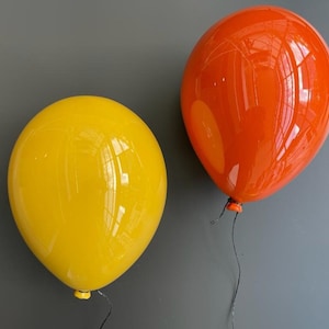 May include: Two balloon-shaped wall decorations. One is bright yellow, the other is orange. Each has a black string and a small yellow knot at the bottom. The balloons are glossy and appear to be made of glass or a similar material.