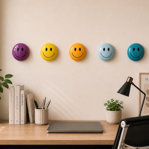 Peut inclure: Cinq décorations murales smiley face colorées en violet, jaune, orange, bleu clair et sarcelle. Chacune a une finition brillante et une expression joyeuse. Elles sont montées sur un mur neutre au-dessus d'un bureau avec des fournitures de bureau et un ordinateur portable.