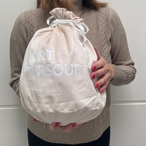 May include: A soft, light beige velvet drawstring pouch featuring a white ribbon closure at the top. The front of the bag displays the white printed text ART ARSOUF in a clean, sans-serif font. The pouch has a rounded, flexible shape.