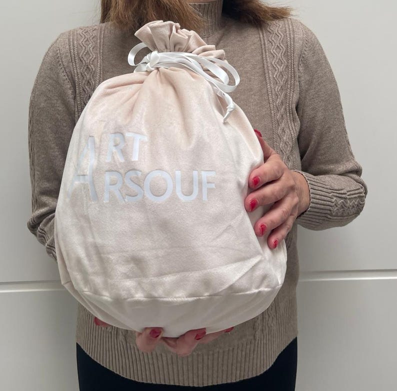 May include: A soft, light beige velvet drawstring pouch featuring a white ribbon closure at the top. The front of the bag displays the white printed text ART ARSOUF in a clean, sans-serif font. The pouch has a rounded, flexible shape.