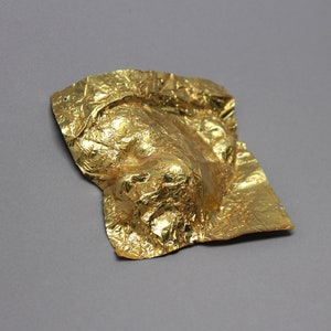 May include: A gold foil mask with a wrinkled texture. The mask is shaped like a face and has a three-dimensional appearance.