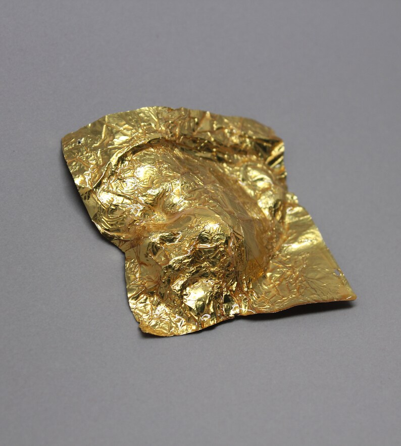 May include: A gold foil mask with a wrinkled texture. The mask is shaped like a face and has a three-dimensional appearance.