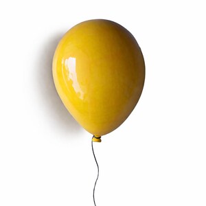 Puede incluir: Un objeto con forma de globo de color amarillo brillante y acabado lustroso con una superficie lisa y reflectante. El globo presenta un peque&ntilde;o nudo oscuro en la base, de donde cuelga un hilo negro fino, creando una imagen simple y limpia.