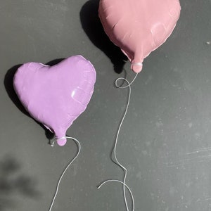 Op de afbeelding: Twee hartvormige ballonnen, &eacute;&eacute;n roze en &eacute;&eacute;n paars, zijn aan elkaar gebonden met een wit touwtje. De ballonnen zweven tegen een grijze achtergrond.