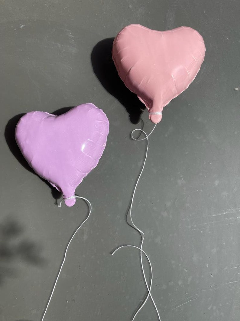 Op de afbeelding: Twee hartvormige ballonnen, &eacute;&eacute;n roze en &eacute;&eacute;n paars, zijn aan elkaar gebonden met een wit touwtje. De ballonnen zweven tegen een grijze achtergrond.