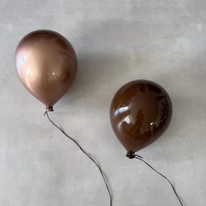 Puede incluir: Dos globos marrones con cuerdas negras sobre un fondo neutro. Un globo es de color bronce metálico, y el otro es marrón chocolate oscuro. Los globos son brillantes y parecen estar flotando.