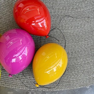 Puede incluir: Tres objetos decorativos lisos y brillantes con forma de globo en rojo vibrante, rosa intenso y amarillo soleado. Cada globo tiene un hilo negro fino unido al nudo inferior, creando la apariencia realista de globos de helio flotantes.