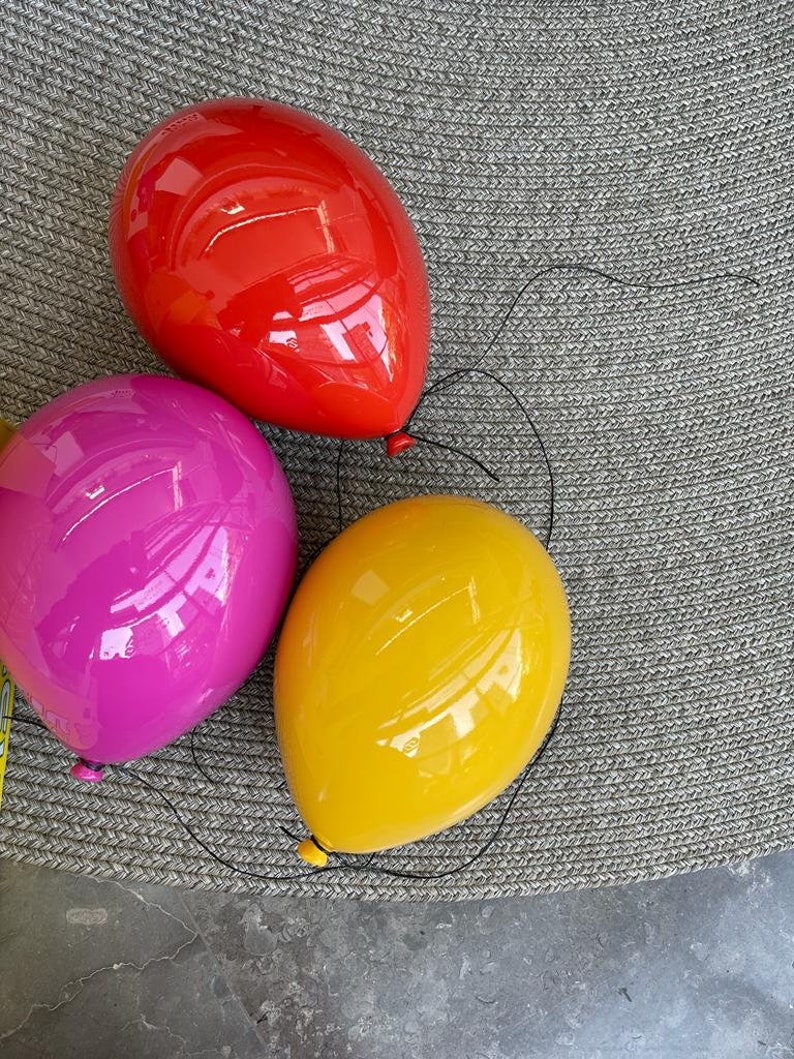 Puede incluir: Tres objetos decorativos lisos y brillantes con forma de globo en rojo vibrante, rosa intenso y amarillo soleado. Cada globo tiene un hilo negro fino unido al nudo inferior, creando la apariencia realista de globos de helio flotantes.