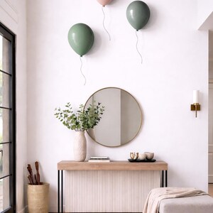 K&ouml;nnte beinhalten: Ein modernes Interieur mit drei Wanddekorationen in Form von Ballons in Rosa und Gr&uuml;n. Ein runder Spiegel h&auml;ngt &uuml;ber einer Konsole mit einer Vase mit Gr&uuml;npflanzen. Ein neutraler Teppich und eine Ottomane vervollst&auml;ndigen den Raum.