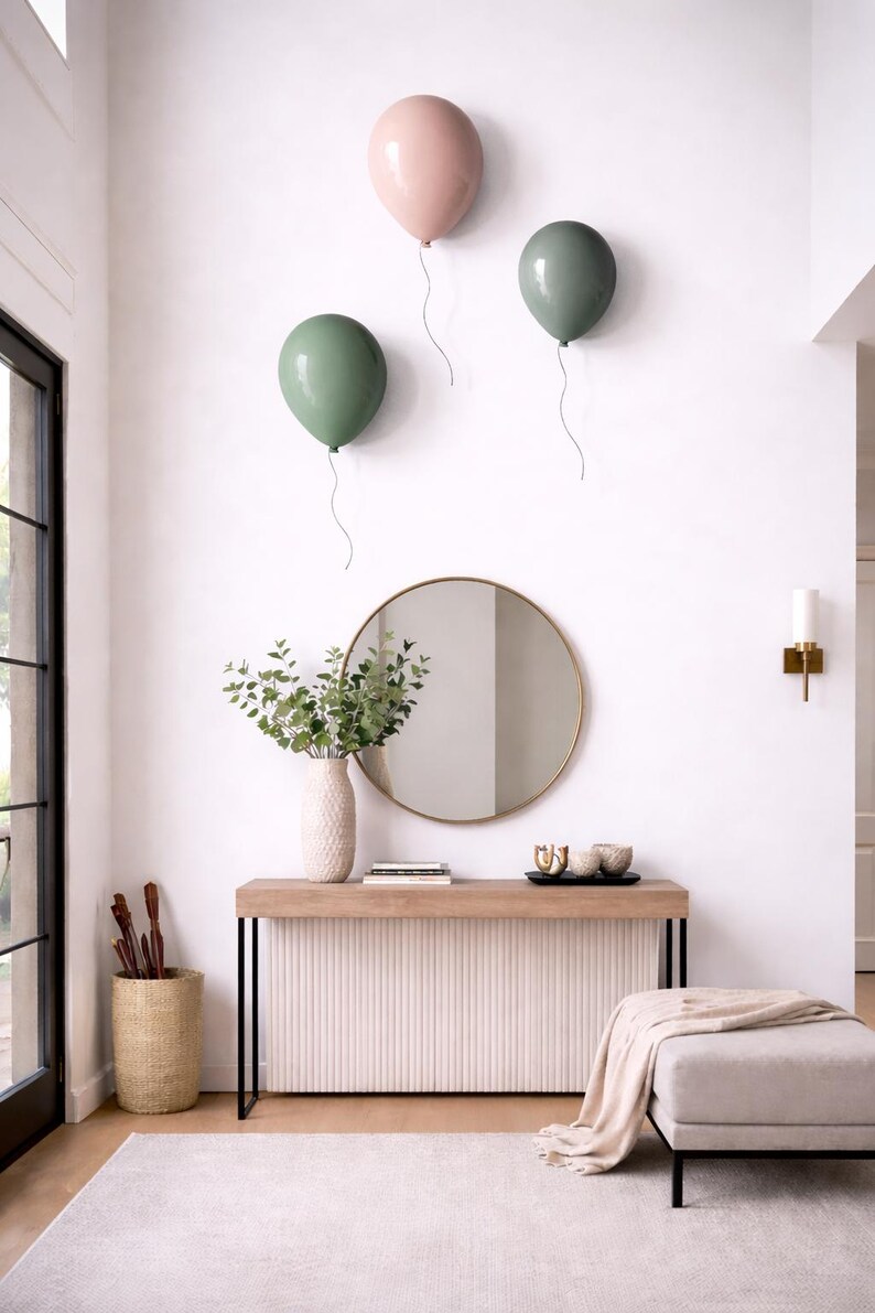 K&ouml;nnte beinhalten: Ein modernes Interieur mit drei Wanddekorationen in Form von Ballons in Rosa und Gr&uuml;n. Ein runder Spiegel h&auml;ngt &uuml;ber einer Konsole mit einer Vase mit Gr&uuml;npflanzen. Ein neutraler Teppich und eine Ottomane vervollst&auml;ndigen den Raum.